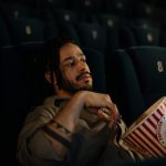 Man sitting alone in a movie theater, holding a large bucket of popcorn.